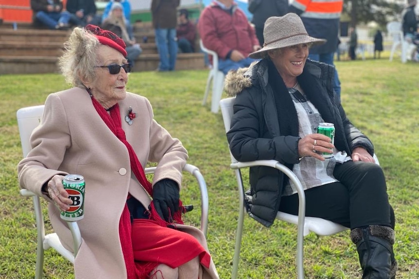 Two women drinking cans of VB and sitting in deck chairs on a lawn.