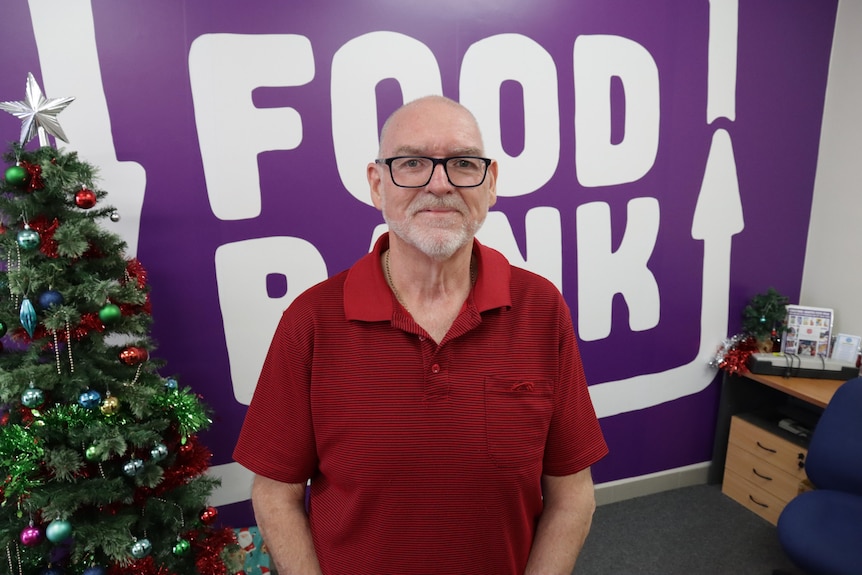 A bald man with black-rimmed glasses and a red shirt standing in front of a wall with the Foodbank logo on it
