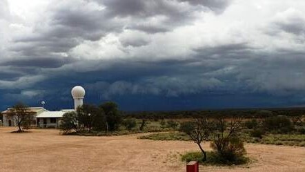 A wide shot of a small, white weather station. Bushland surrounds and dark clouds hang overhead.