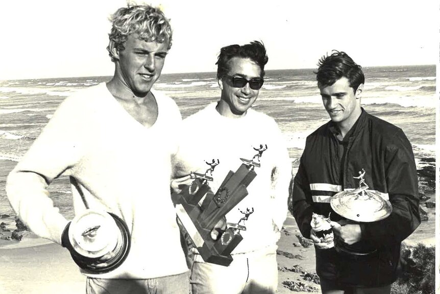 Three men stand in front of a surf break holding trophies.