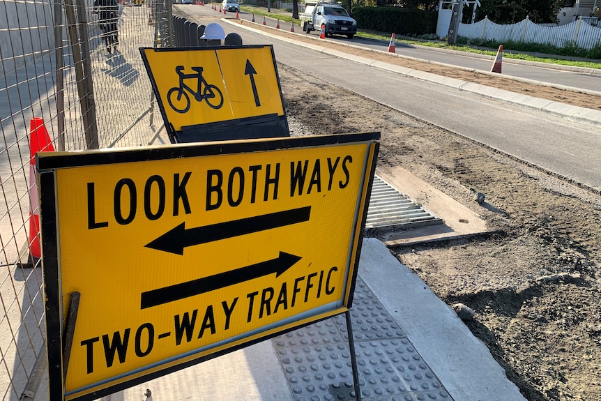 A yellow and black 'Look Both Ways - Two-Way Traffic' sign by the side of a dug-up footpath and road.