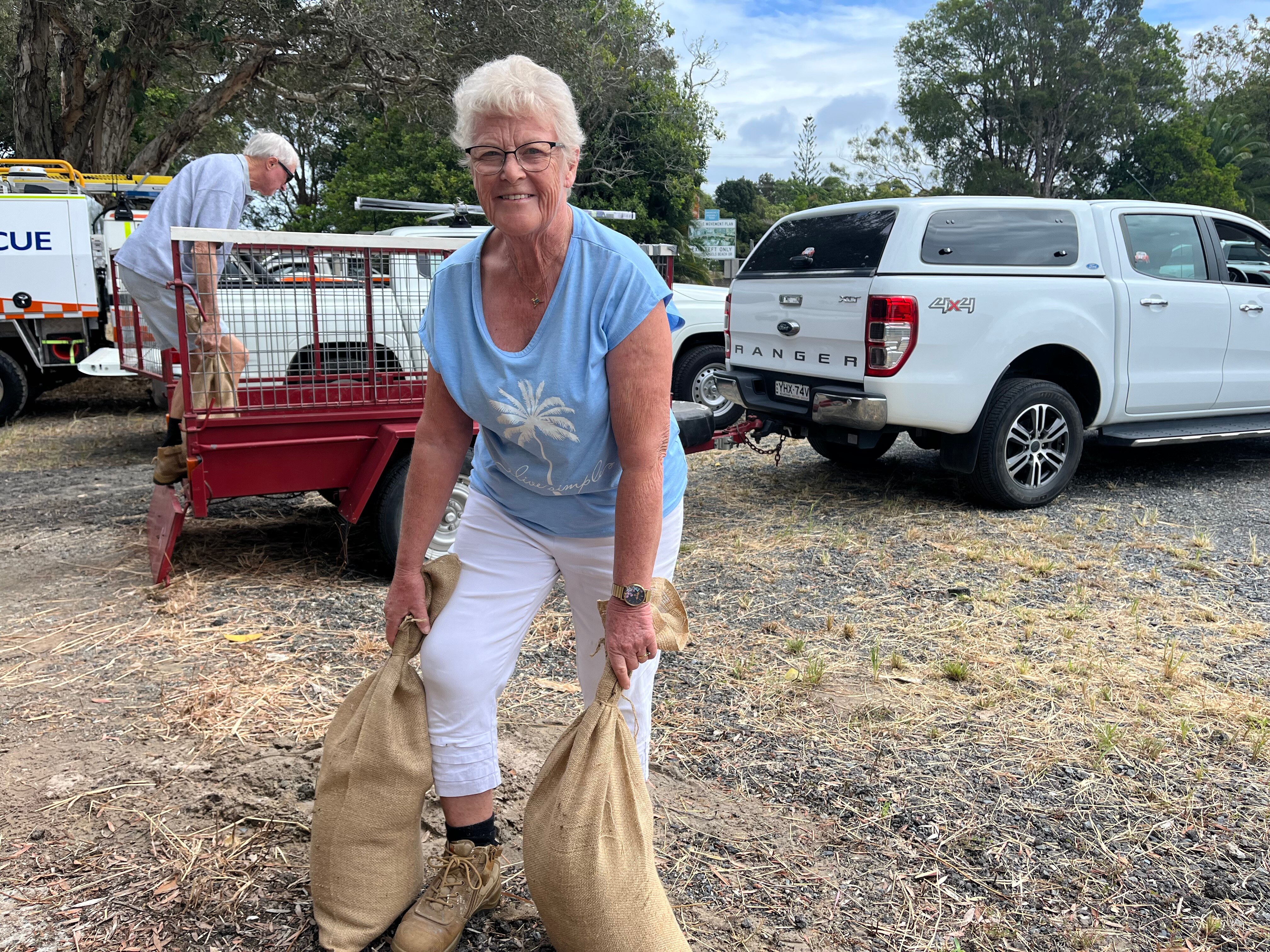 An older woman in a blue shirt, lifting two sandbags with cars in the background.