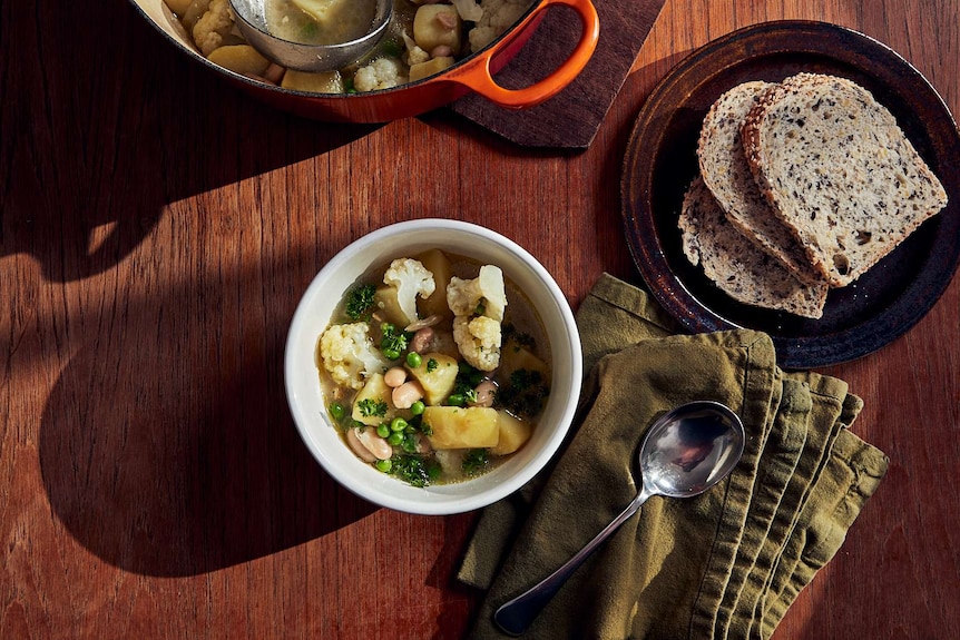 A bowl of vegetable soup sits beside a napkin, spoon, plate of bread and dutch oven of soup, a warming dinner.