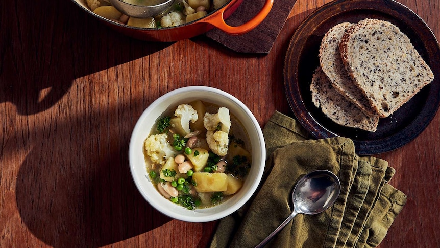 A bowl of vegetable soup sits beside a napkin, spoon, plate of bread and pot of soup, a warming dinner recipe.