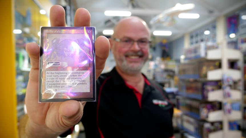 A man holding a shiny trading card is smiling, there is a store with stocked shelves behind him