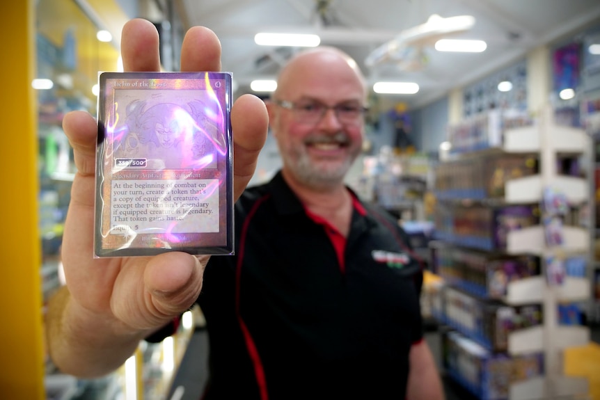 A man holding a shiny trading card is smiling, there is a store with stocked shelves behind him