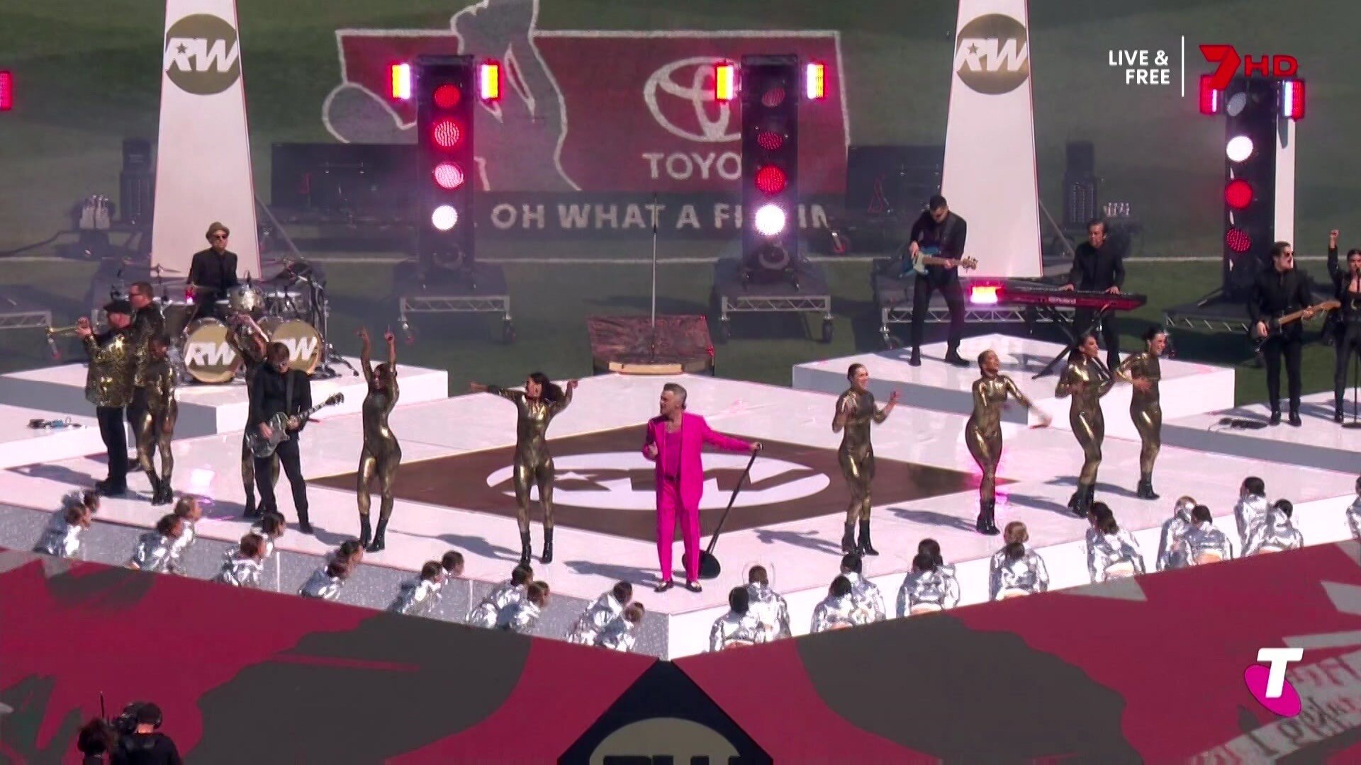 Robbie Williams dances with performers in shiny silver costumes on stage.