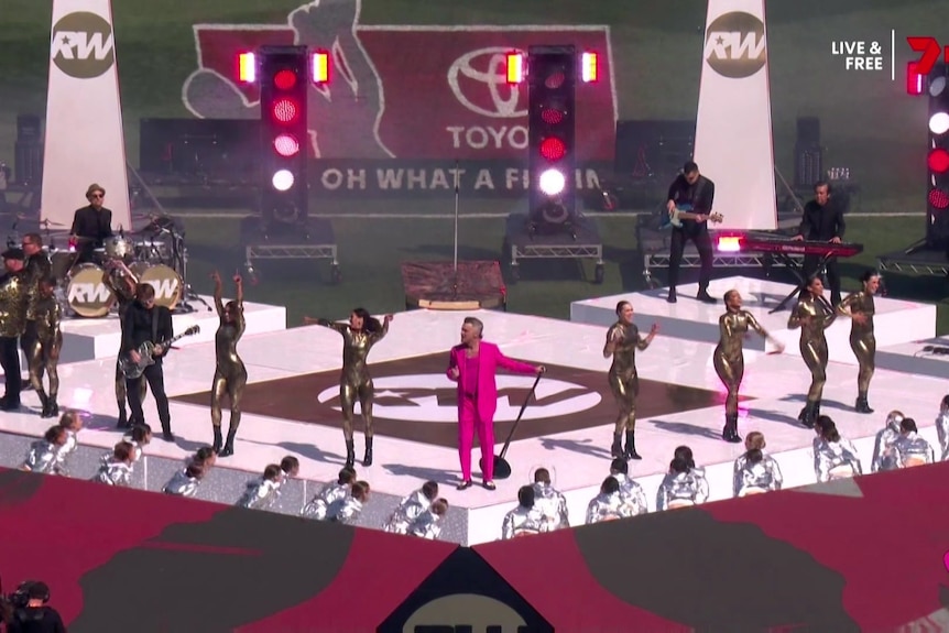 Robbie Williams dances on a stage with shiny silver costumed performers.