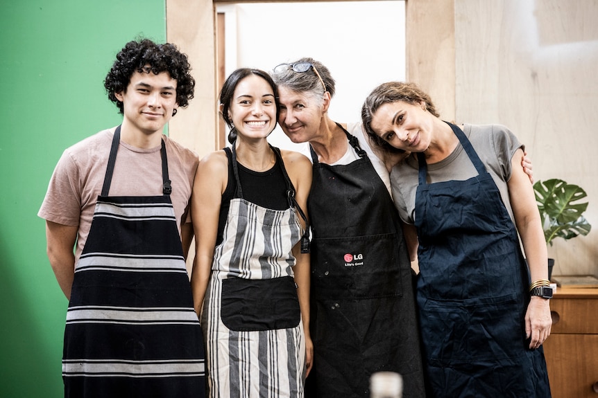 Two young people, and two middle-aged women, stand closely together, wearing aprons, arms around each other