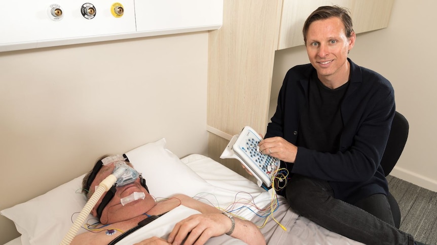 A professor holding a control pad with wires attached to a patient wearing a CPAP mask undergoing a study