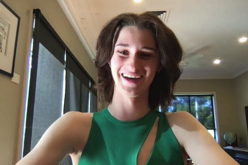 A teenager with shoulder-length hair, wearing a sleeveless green top smiles at the camera