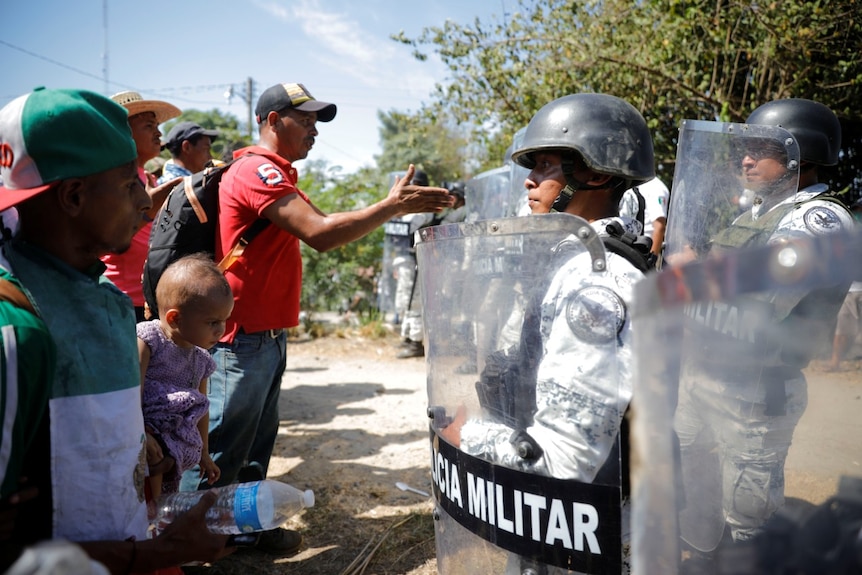 a man holds out his arm as he speaks to mexican national guard members in riot gear lined up near trees