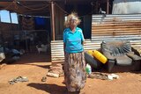 An elderly Indigenous woman in a blue shirt stands out the front of a corrugated iron building
