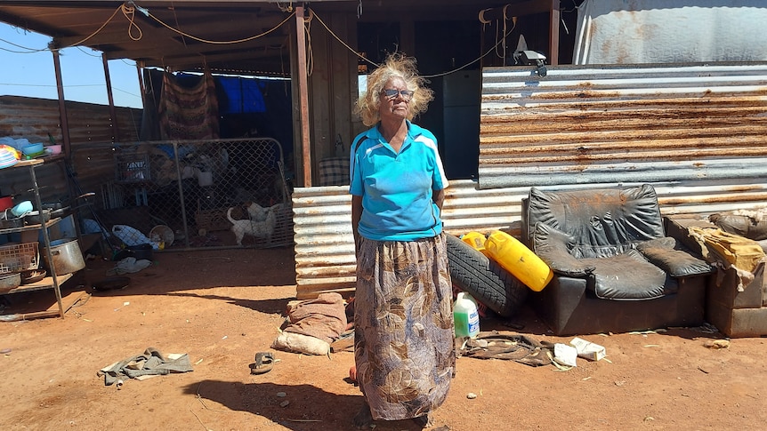 An elderly Indigenous woman in a blue shirt stands out the front of a corrugated iron building