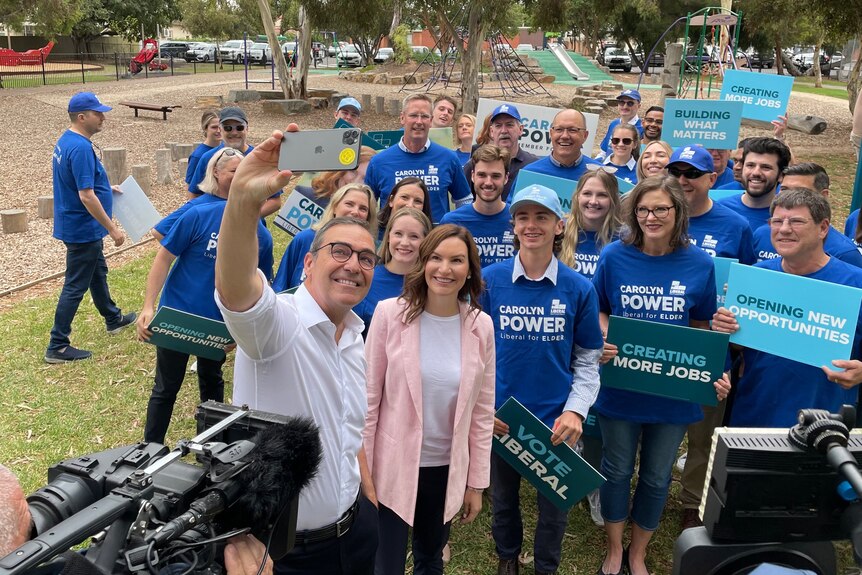 A man holds up a camera for a selfie in a park with a woman and a group of supporters wearing blue campaign tshirts