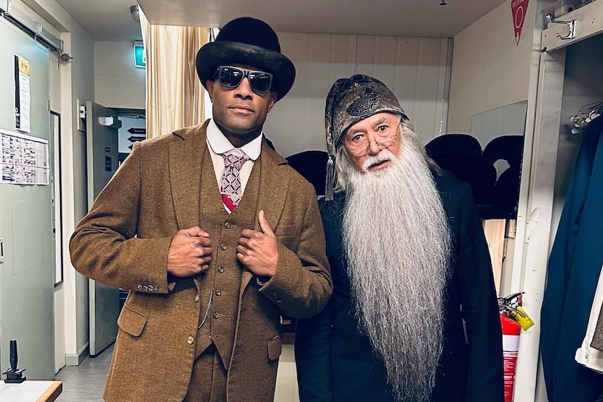 Two men, one a Torres Strait Islander man in brown jacket and bowler cap, the other a long-bearded wizard.