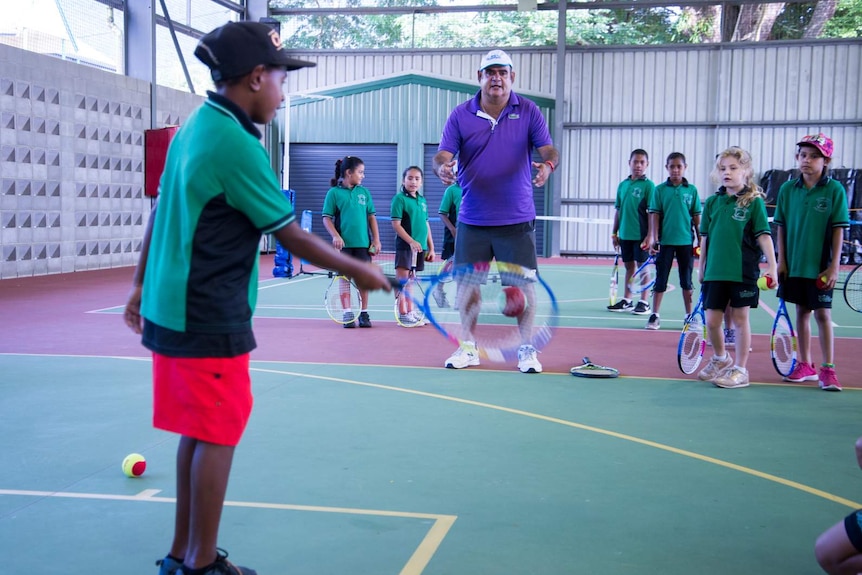 A school student hits a tennis ball back to Ian Goolagong during a racquet drill.