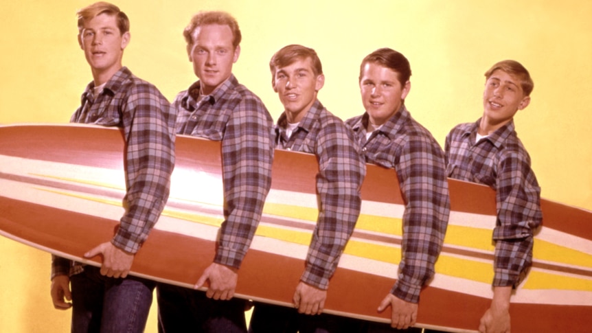 Brian Wilson, Mike Love, Dennis Wilson, Carl Wilson, David Marks from band The Beach Boys pose with a surfboard