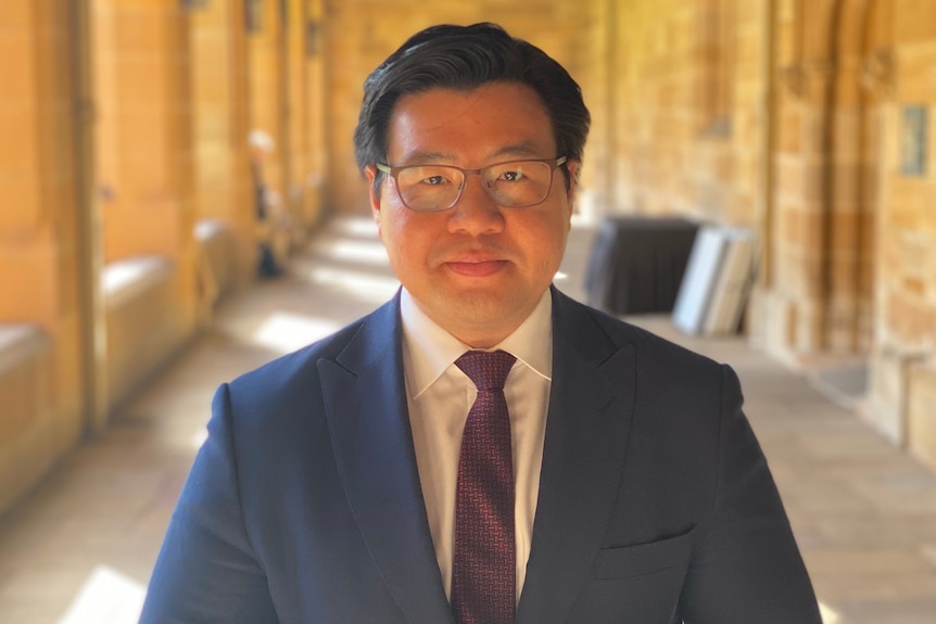 A man in a suit poses for a photo in what looks like a hall of a sandstone university