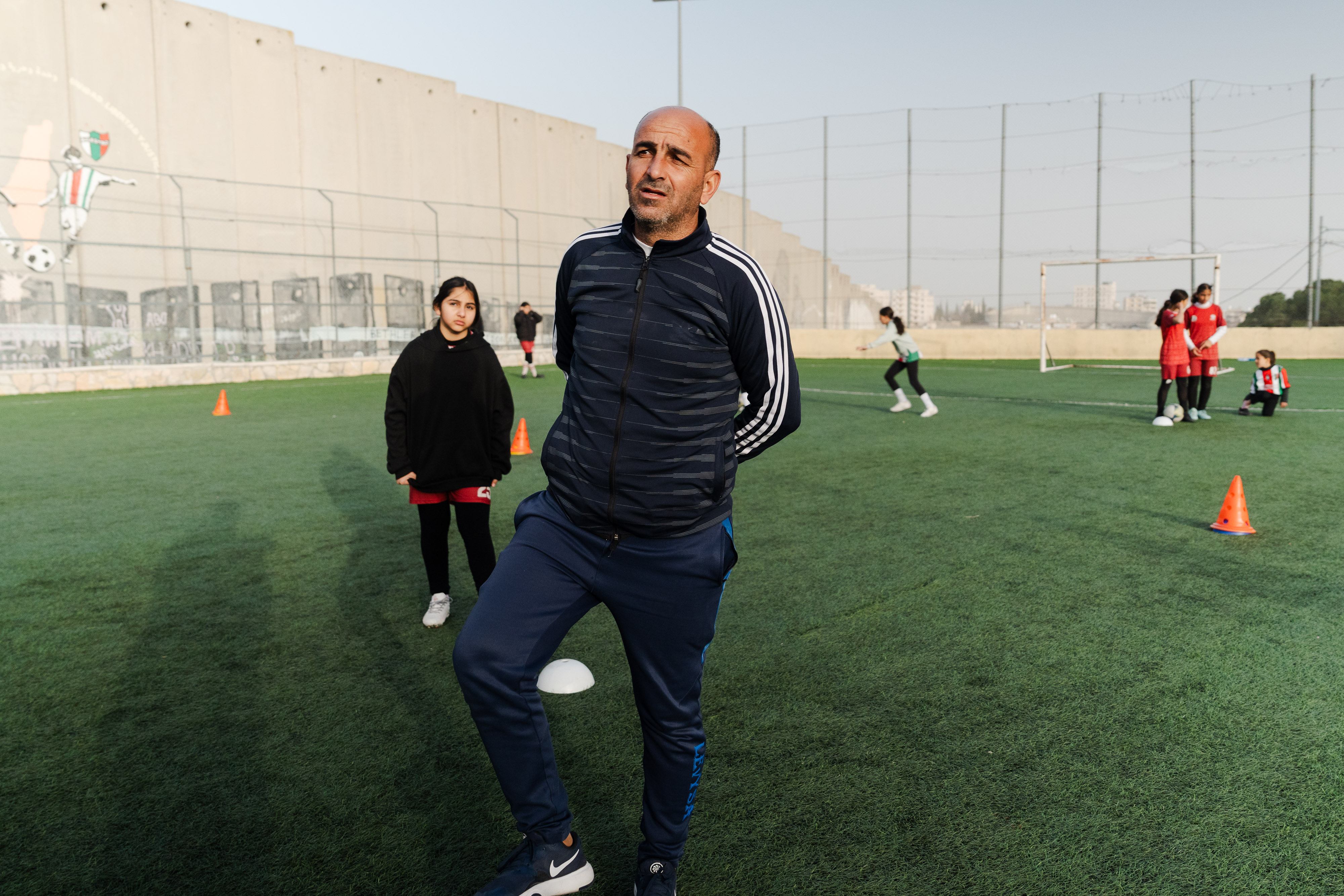 Un palestino con suéter y jeans está en un campo de fútbol con otros jugadores.