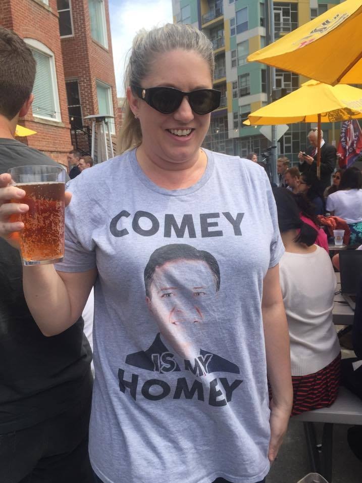 Angela wears a grey t-shirt that reads 'Comey is my Homey' while holding a pint of beer.