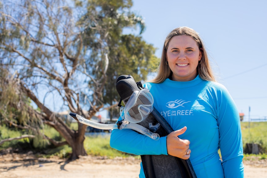 A smiling woman with long blonde hair, wearing a rash shirt and clutching a snorkel and flippers.