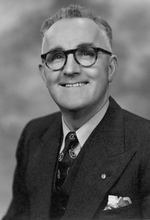 A man with round glasses and wearing a suit smiles at the camera in an old black and white photo