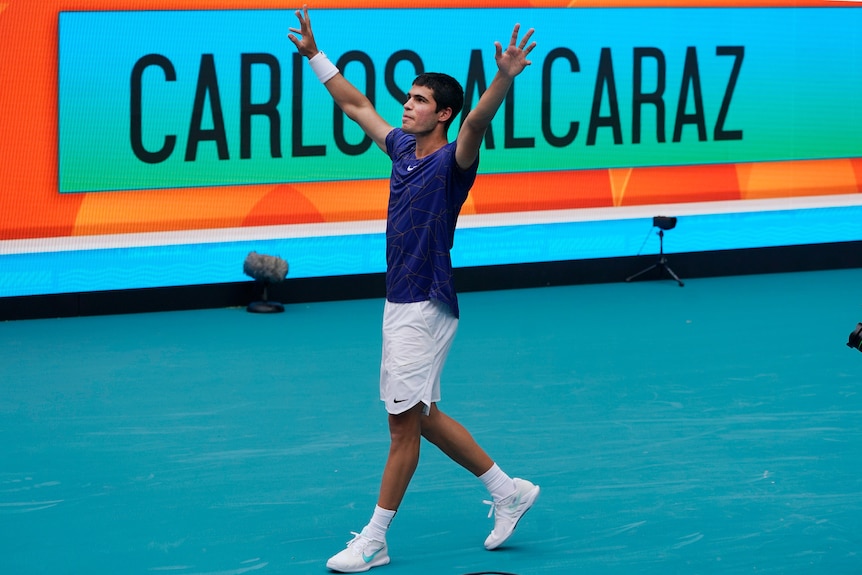 Spanish tennis star Carlos Alcaraz raises his arms in triumph as he walks with a 'Carlos Alcaraz' sign behind him. 