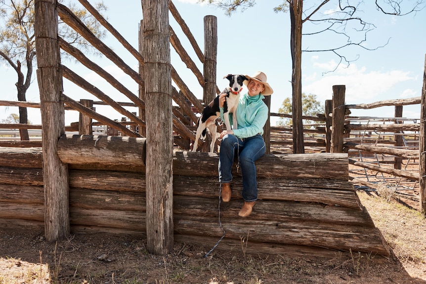A dog stands near a woman as she sits on an old wooden cattle loading ramp.