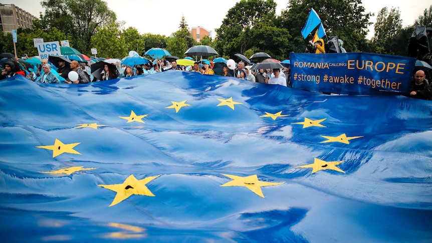A large EU flag floats in the foreground as Romanian crowds hold umbrellas in the distance.