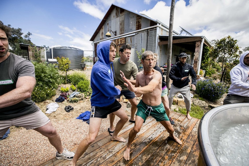 A group of men in swimmers stand with legs apart and move their arms