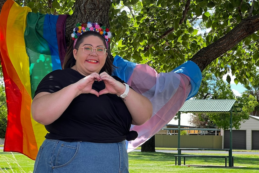 A woman poses in front of a tree draped with the rainbow gay pride flag and the pink, bue and white transgender flags