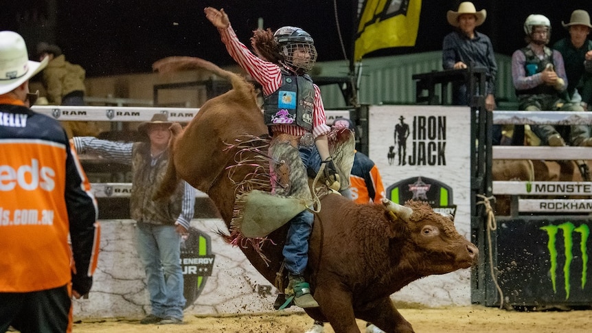 Caydence Fouracre riding an orange bucking bull in a rodeo arena.