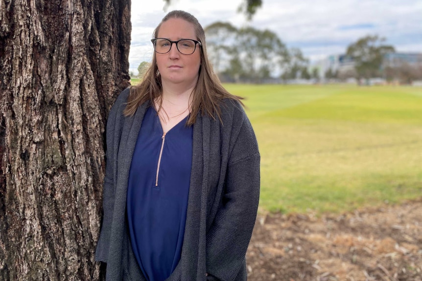 A woman stands in a grey jumper and blue shirt against a tree looking sombre at the camera