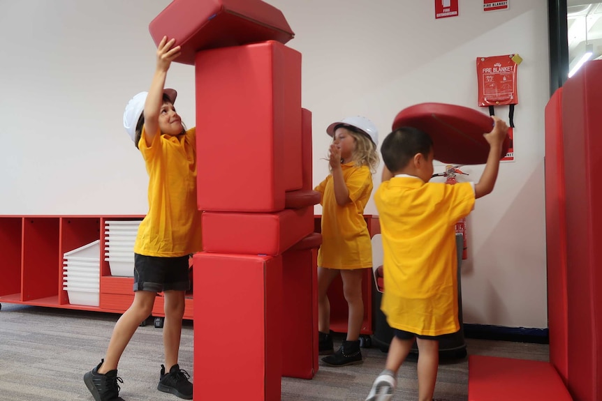 Three children stack large red building blocks