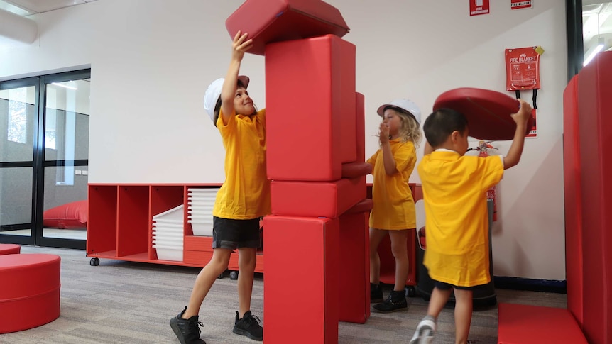 Three children stack large red building blocks