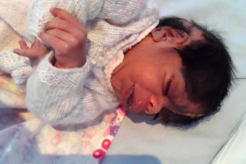 An unwell newborn girl lies in a hospital cot wearing a light pink woollen jumper.