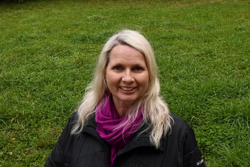 A woman with long blonde hair smiles at the camera with grass in the background.