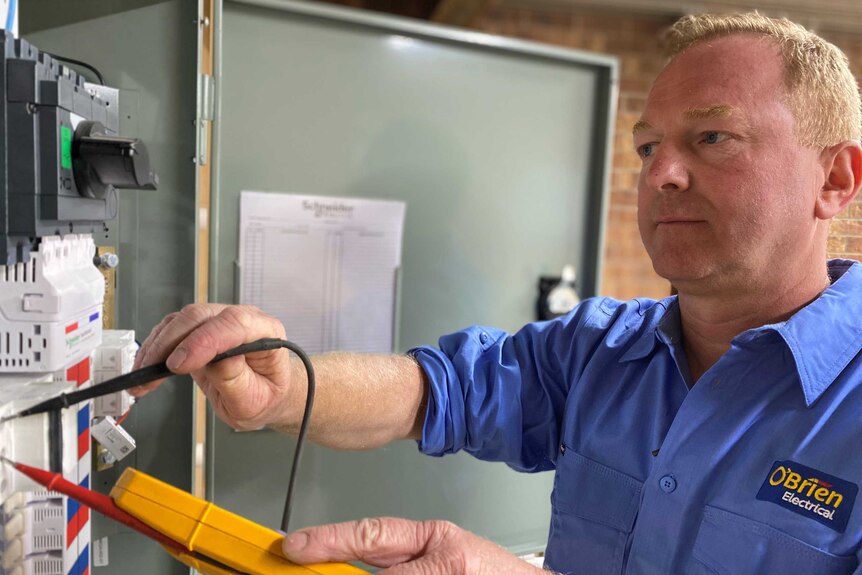 A male electrician wearing a blue shirt holds a yellow power reading device against a meter board.