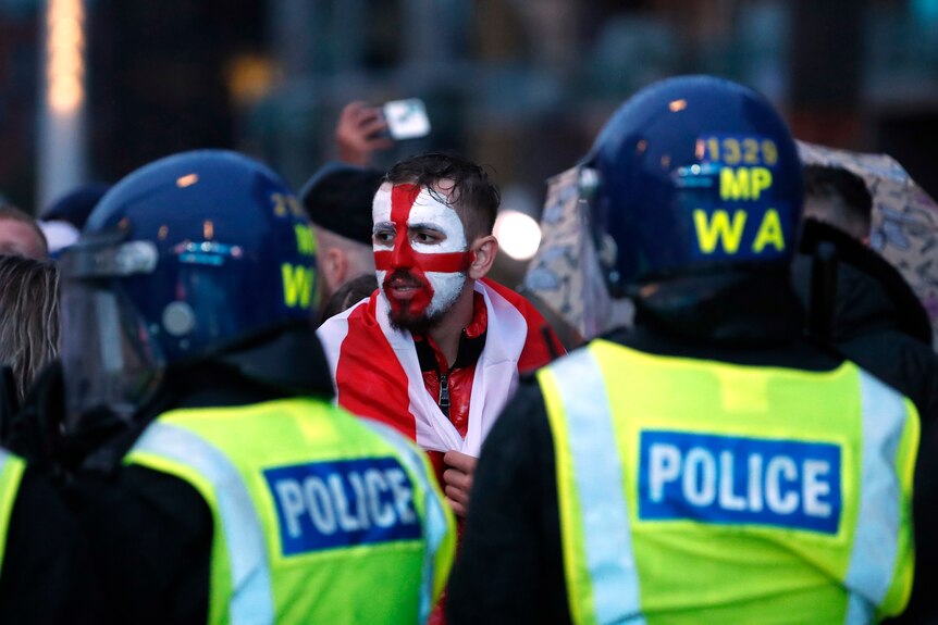 Two London police officers in the fore-ground with their backs turned, while a fan with English flag face-paint looks on