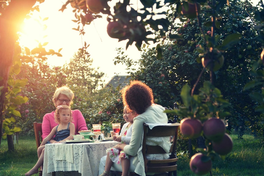 Two women and two children sit in an orchard at a table smiling over food and drink.
