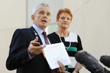 Malcolm Roberts holds a document and gestures. Pauline Hanson stands behind him.