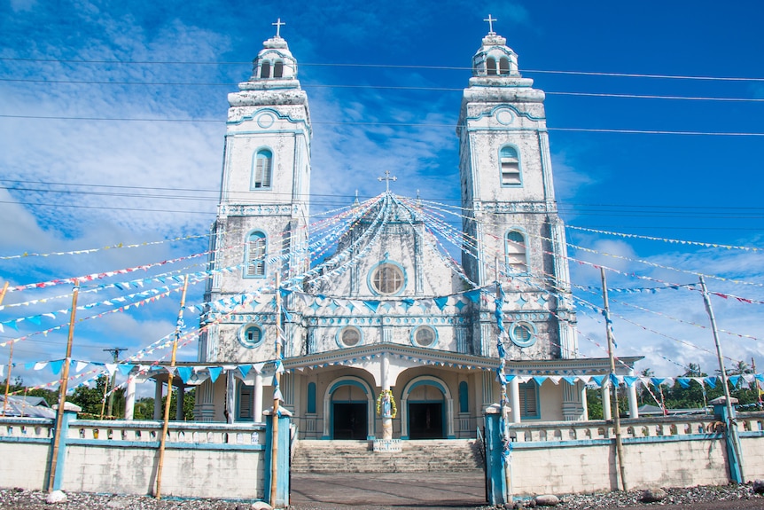 A white church with light blue trimming and two towers, covered in blue and white flags.