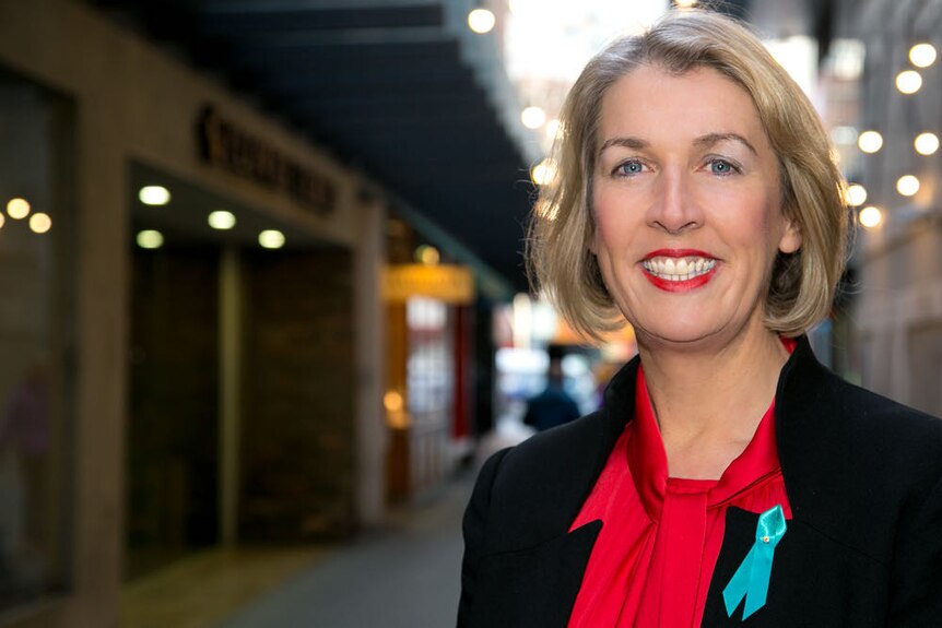 A woman with blonde hair, smiling, wearing red lipstick, red shirt, black jacket, blue eyes, in an alleyway, lights behind.