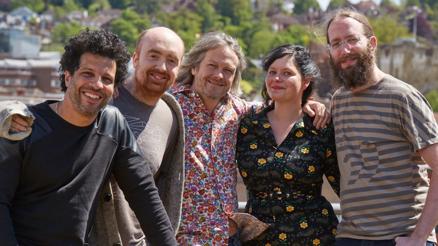 Four men and a woman with their arms around each other smile at the camera on a sunny day.