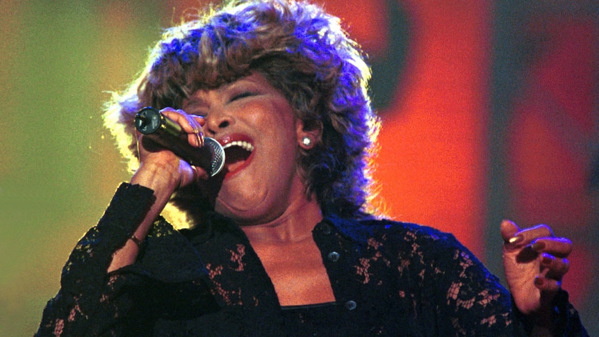 Tina Turner belting into a microphone. She has short, curly blond hair and red lipstick. She's wearing a black lace chemise. 