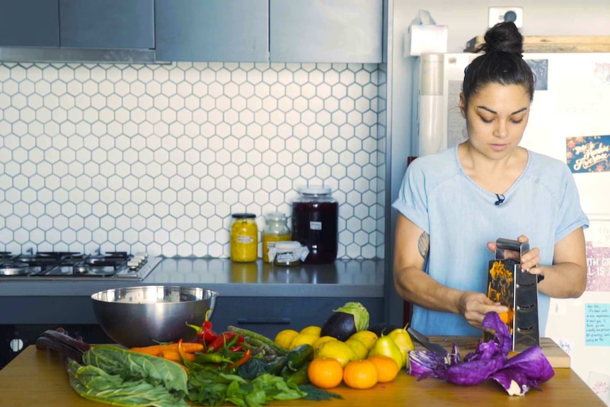 Cass Hay standing in her kitchen grating carrot, with an array of veggies on the bench.