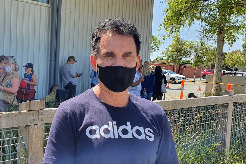 A man with dark hair, a black mask and an Adidas t-shirt stands in front of a fence