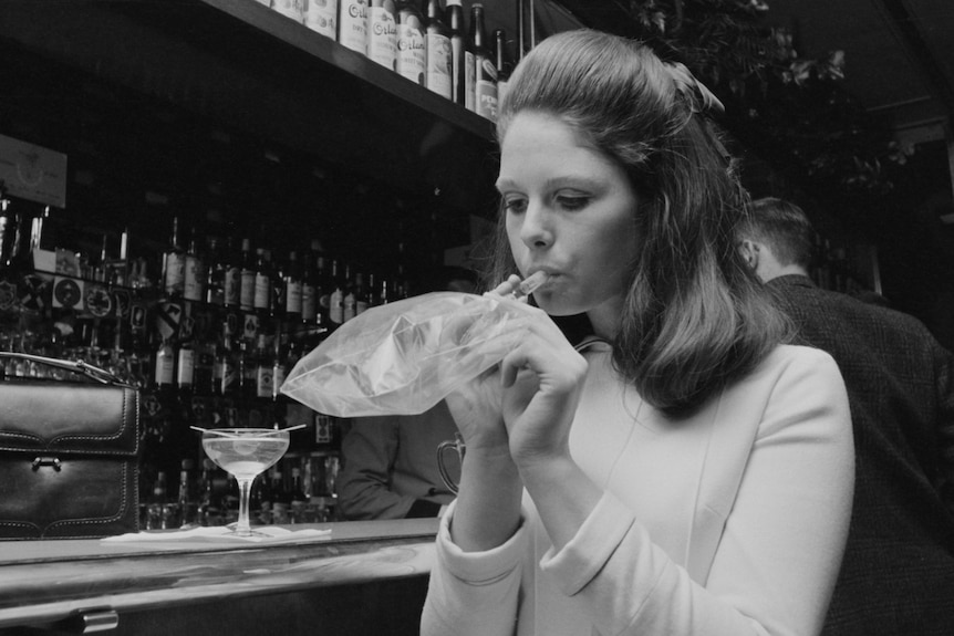 A black and white photo of a young woman at a bar, blowing into a small plastic bag