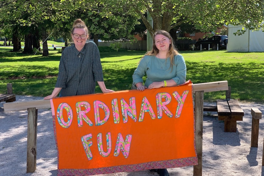 Two grinning young women lean on a wooden post with a large sign on an orange banner with Ordinary Fun written on it.